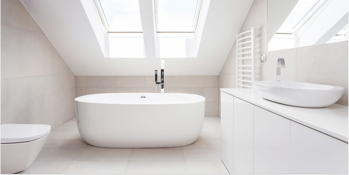 Freistehende weiße Badewanne unter zwei Dachfenstern in modernem Badezimmer mit weißem Waschbecken und Toilette.