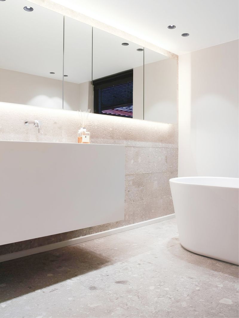 Modernes Badezimmer mit einem eleganten weißen Waschtisch, großen Spiegeln, sanfter Beleuchtung und einer freistehenden Badewanne auf strukturiertem Steinboden.